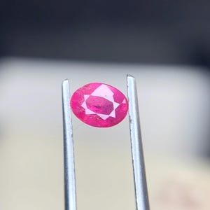 Puede incluir: Una gema de color rojo rosado vibrante y corte ovalado, sostenida firmemente entre las dos puntas metálicas de unas pinzas plateadas. La piedra presenta una superficie facetada que refleja la luz, creando un patrón geométrico distintivo.