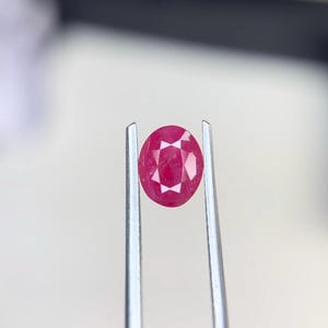 Puede incluir: Una piedra preciosa de color rosa rojizo vibrante y corte ovalado, sostenida por unas pinzas de metal plateado. La piedra presenta una superficie facetada con un patrón distintivo en forma de estrella que refleja la luz desde su centro.