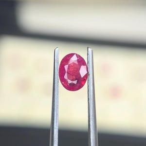 Puede incluir: Una gema de rubí de corte ovalado con una superficie facetada, que muestra un color rojo intenso. La piedra preciosa se sostiene de forma segura entre las dos puntas metálicas de unas pinzas plateadas, resaltando su claridad y corte.
