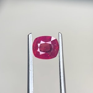 Puede incluir: Una gema roja de talla cojín sostenida por unas pinzas metálicas de tono plateado. La piedra presenta una superficie facetada que refleja la luz, mostrando un tono carmesí profundo y saturado con una claridad interna muy definida.