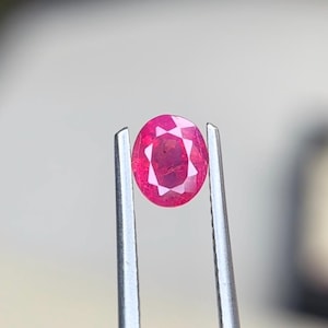 Puede incluir: Una gema de rubí de corte ovalado con una superficie facetada que refleja la luz. La piedra de color rojo rosado intenso se sostiene de forma segura entre las dos puntas metálicas de unas pinzas plateadas, mostrando su claridad y color.