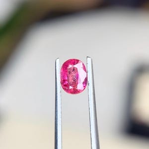Puede incluir: Una gema rosa de corte ovalado, sostenida firmemente por unas pinzas metálicas de tono plateado. La piedra presenta una superficie facetada que refleja la luz, mostrando un tono magenta profundo y saturado con claridad interna visible.