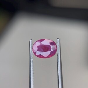 Puede incluir: Una piedra preciosa de color rosa vibrante con corte ovalado, sostenida por unas pinzas de metal plateado. La piedra presenta una superficie facetada que refleja la luz, mostrando una calidad translúcida y un tono magenta profundo en su forma.