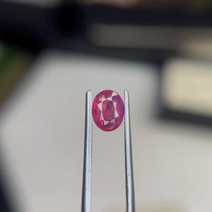 Puede incluir: Una gema de color rojo rosado con talla ovalada, sostenida firmemente por unas pinzas de metal plateado. La piedra facetada muestra reflejos de luz internos y una superficie clara y pulida, destacando su forma ovalada distintiva.