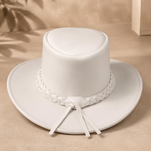 May include: A solid white hat with a flat top and a wide brim. The hat features a decorative braided band around the base of the crown, which includes two hanging strips of material at the front, creating a clean and uniform appearance.
