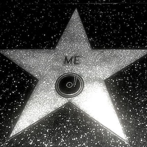 May include: A five-pointed star embedded in a dark, speckled ground. The star is light-colored with a metallic texture. The word ME is printed in black capital letters above a circular black icon featuring a stylized record and an exclamation point.
