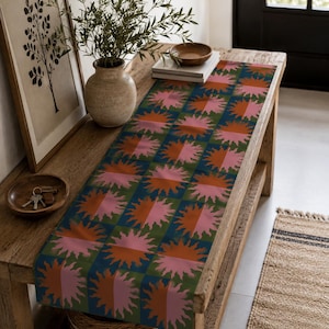 Puede incluir: Un camino de mesa rectangular con un patrón de cuadrícula repetitivo de motivos de rayos de sol. Cada cuadrado alterna entre fondos azul oscuro y verde oliva, con cada sol dividido en dos mitades de colores rosa y naranja contrastantes.