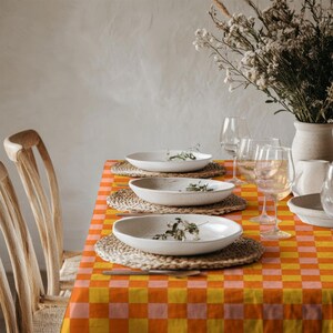 May include: A table setting featuring a vibrant orange and yellow checkered tablecloth, three speckled white ceramic bowls placed on woven round placemats, clear glass stemware, and a vase filled with dried white flowers on a wooden table.