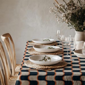 Può includere: Una tovaglia rettangolare con un motivo ripetuto di archi intrecciati blu navy scuro e arancione bruciato su sfondo crema. Tre ciotole in ceramica bianco sporco macchiate sono posizionate su tovagliette rotonde in vimini intrecciato.