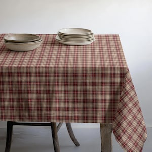 May include: A rectangular tablecloth featuring a plaid pattern in shades of red, dark brown, and beige. A stack of five white ceramic plates sits on the left side, and a single white ceramic bowl is placed on the right side of the table.