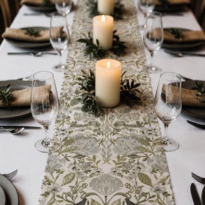 May include: A long rectangular table runner featuring a repeating botanical pattern in shades of sage green, olive, and grey on a cream background. The runner is centered on a white tablecloth with four lit pillar candles and clear glassware.