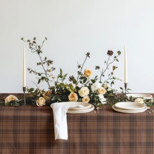 May include: A brown plaid tablecloth set with two white taper candles in metal holders, a floral centerpiece of cream roses and green foliage, and two white ceramic plates with a folded white linen napkin draped over the edge of the table.
