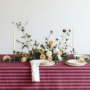 May include: A table setting featuring a dark pink and light pink striped tablecloth, a central floral arrangement of cream and peach roses with green foliage, two tall cream taper candles in holders, and white ceramic plates with a white napkin.
