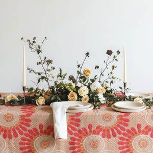 Könnte beinhalten: Ein Tischgedeck mit einer Tischdecke mit orangefarbenem und rosafarbenem Kreismuster, einem Blumenarrangement aus cremefarbenen Rosen und Grünpflanzen, zwei hohen weißen Stabkerzen in dunklen Haltern sowie weißen Keramiktellern mit Serviette.