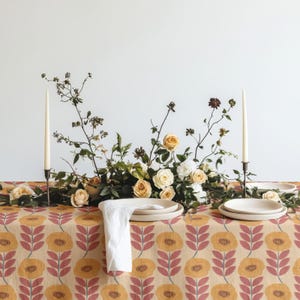 May include: A table setting featuring a tablecloth with a repeating yellow and pink floral pattern, topped with a centrepiece of white and peach roses, two tall cream taper candles in dark holders, and white ceramic plates with a white napkin.