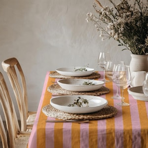 May include: A table setting featuring three white ceramic bowls placed on round woven placemats atop a mustard yellow and pink striped tablecloth. Clear glass stemware and a ceramic vase filled with dried flowers are arranged on the table.