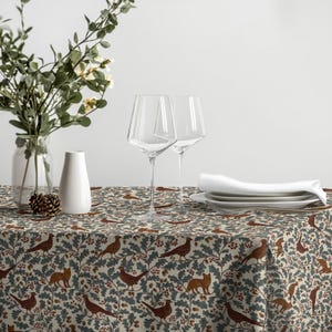 Könnte beinhalten: Eine Tischdecke mit einem sich wiederholenden Muster aus braunen Füchsen, Vögeln und Eicheln inmitten grüner Eichenblätter auf cremefarbenem Grund. Zwei klare Weingläser, eine weiße Keramikvase und ein Stapel weißer Teller mit Serviette.