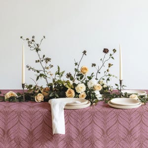 Może przedstawiać: Malwowy obrus w biały wzór paproci ozdobiony jest stroikiem z kremowych róż i zieleni. Dwie wysokie, cienkie, kremowe świece stożkowe umieszczone są w ciemnych metalowych świecznikach, obok znajdują się białe ceramiczne talerze i serwetka.