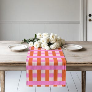May include: A rectangular table runner featuring a vibrant plaid pattern of alternating orange, light pink, and white vertical and horizontal stripes. A bouquet of white roses rests on the centre of the runner between two white ceramic plates.