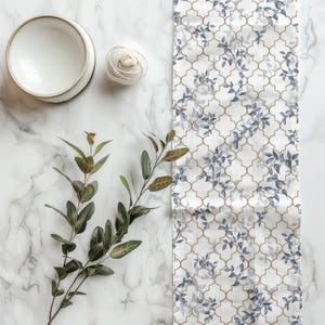 May include: A white ceramic bowl, a small ball of white yarn, a green eucalyptus branch, and a long fabric table runner featuring a white background with a gold quatrefoil lattice pattern and blue leaf motifs arranged in a repeating design.