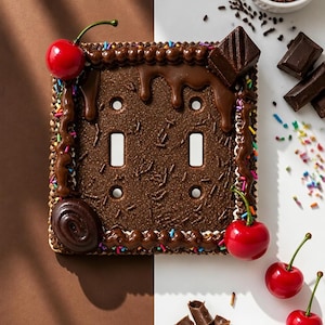 May include: A square double light switch cover plate decorated to resemble a chocolate dessert. The brown surface features chocolate icing, colorful sprinkles, two red cherries, a chocolate square, and a swirl-shaped chocolate piece.