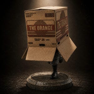 Könnte beinhalten: Eine Figur mit einem Pappkarton, der mit THE ORANGE sowie Gourmet Quality Organic und 26 LBS/IN beschriftet ist. Der Karton wird auf dem Kopf einer Figur mit dunklen Stiefeln und Hose getragen, die auf einem runden, steinartigen Sockel steht.