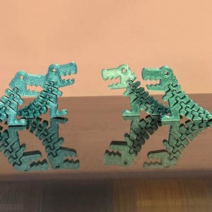 Könnte beinhalten: Zwei bewegliche grüne Dinosaurierfiguren mit segmentierten Körpern und Schwänzen. Jede Figur hat eine strukturierte Oberfläche, ein weit geöffnetes Maul mit sichtbaren Zähnen und ein kleines kreisförmiges Loch oben am Kopf zur Befestigung.