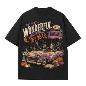 Op de afbeelding: Een zwart t-shirt met een afbeelding van twee skeletten in een paarse cabriolet. De tekst luidt THE MOST WONDERFUL TIME OF THE YEAR en HAUNTED BOOKSTORE NEXT EXIT. Het ontwerp bevat pompoenen, een stapel boeken en een halve maan.