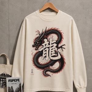 May include: A cream-coloured long-sleeve crewneck sweatshirt featuring a large graphic of a black dragon with red accents. The design includes the Japanese kanji for dragon, the text "日本" and "JAPAN" in small print, and a ribbed collar and cuffs.