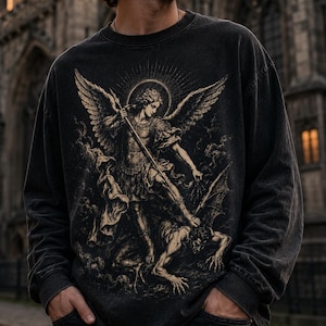 Puede incluir: Una sudadera de manga larga en color gris carbón con un estampado gráfico detallado de un ángel con alas de pie sobre una figura caída. La ilustración está representada en tonos beige y negro, mostrando una escena clásica con una lanza.