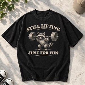 Puede incluir: Una camiseta negra de manga corta con un gráfico de un mapache levantando una barra de pesas. El texto "STILL LIFTING" está impreso en color crema sobre el mapache, y el texto "JUST FOR FUN" está impreso en el mismo color debajo de él.
