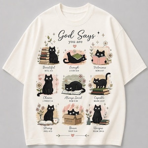 May include: An off-white short-sleeve t-shirt featuring a graphic print with nine illustrations of black cats. The text reads God Says you are, with labels including Beautiful, Enough, Victorious, Chosen, Always Loved, Capable, Strong, Brave, and Unique.