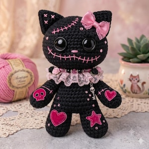 May include: A black crocheted cat doll featuring large black button eyes, pink stitched mouth details, and a pink satin bow on one ear. The doll wears a pink lace collar with beads and a small skull charm, decorated with pink heart and star patches.