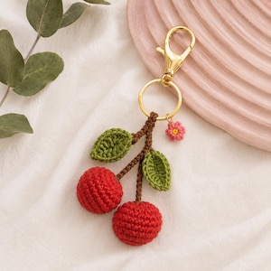 May include: A crochet keyring featuring two round red cherries with brown stems and two green leaves, attached to a gold-toned metal lobster clasp and keyring, accented by a small pink flower charm with a yellow centre.