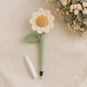 May include: A ballpoint pen featuring a crocheted cover in light green, topped with a crocheted daisy flower with white petals and a yellow center. A small crocheted leaf is attached to the side of the pen, and a separate white plastic pen cap is placed next to it.