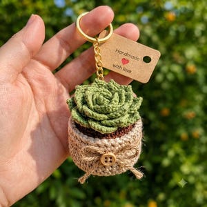 Pode incluir: Um porta-chaves de suculenta em croché com uma planta verde num vaso bege, decorado com um pequeno botão de madeira e um laço de corda. Inclui um anel de metal dourado e uma etiqueta de papel castanho com a inscrição Handmade e um coração vermelho.