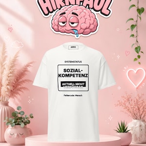 Puede incluir: Una camiseta blanca de manga corta con un gráfico rectangular negro con el texto SYSTEMSTATUS, SOZIAL-KOMPETENZ, AKTUELL NICHT VERFÜGBAR y Fehlercode: Mensch. Sobre la camiseta hay un personaje de cerebro de dibujos animados con el texto HIRNFAUL.