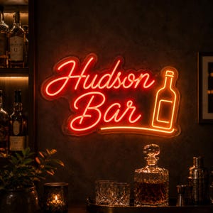 May include: A bright orange neon sign featuring the text Hudson Bar in a cursive script font, accompanied by a simple outline of a bottle. The sign is mounted on a dark wall, emitting a warm glow that illuminates the surrounding bar area.