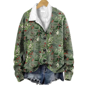 Könnte beinhalten: Eine langärmelige, geknöpfte Strickjacke in gedämpftem Salbeigrün mit einem botanischen Druck aus Stechpalmenblättern, roten Beeren und weißen Blüten. Das Kleidungsstück hat eine weiße Hemdschicht darunter und wird mit blauen Jeansshorts kombiniert.