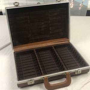 May include: A brown and silver rectangular storage case with a metal frame and a brown handle. The interior features a dark brown velvet lining with three partitioned sections, each containing multiple slots for organized storage.
