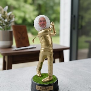 May include: A gold-colored golfer figurine standing on a circular black base with green artificial turf. The golfer has a white golf ball for a head featuring a printed photograph of a smiling person. The base displays the text BEST DAD BY PAR.