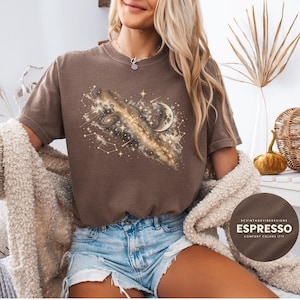May include: A brown short-sleeve t-shirt featuring a graphic print of a galaxy, stars, and a crescent moon. The design includes swirling cosmic clouds, constellations, and sparkling star accents. A circular badge on the image reads KCVINTAGEVIBEDESIGNS ESPRESSO COMFORT COLORS 1717.