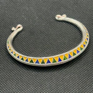 May include: A silver-toned open cuff bracelet featuring a repeating pattern of small triangles filled with blue, yellow, and green enamel. The metal band has a textured, beaded border along the edges and terminates in two small, curled scroll ends.