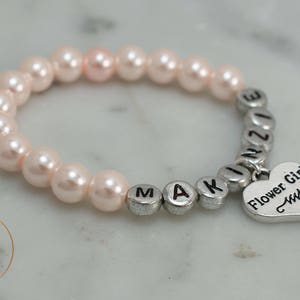 May include: A pink pearl bracelet with silver letter beads spelling "MAKINZIE" and a silver heart charm that says "Flower Girl".