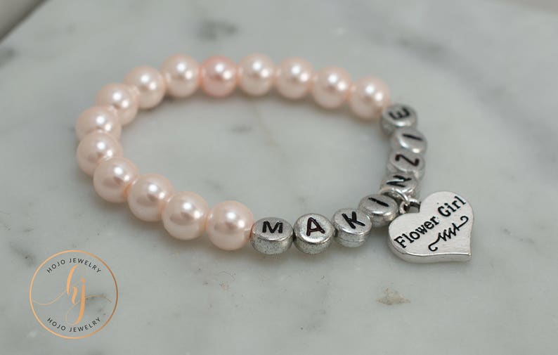 May include: A pink pearl bracelet with silver letter beads spelling "MAKINZIE" and a silver heart charm that says "Flower Girl".