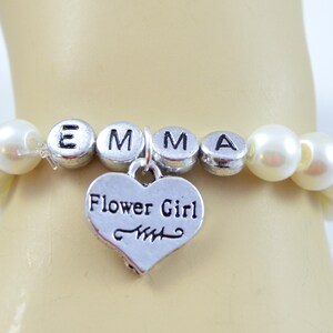 May include: A white pearl bracelet with a silver charm that says "Flower Girl" and silver letter beads that spell "EMMA".