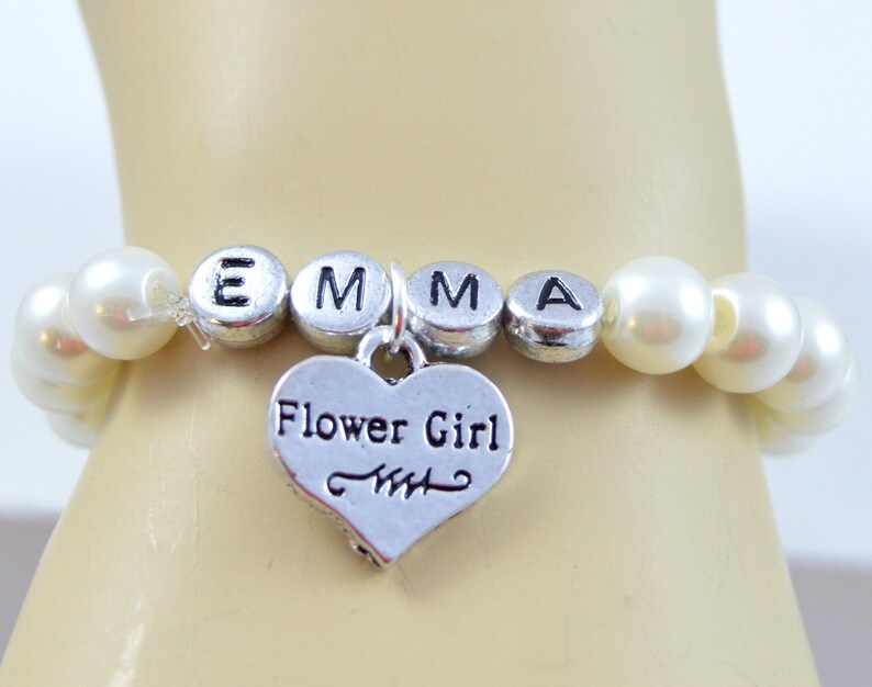May include: A white pearl bracelet with a silver charm that says "Flower Girl" and silver letter beads that spell "EMMA".