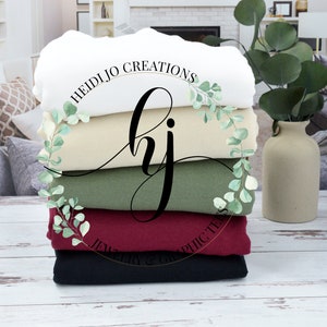 May include: Stack of five blank t-shirts in white, beige, green, burgundy, and black. The shirts are folded and stacked on a white wooden surface. A gold circle with the text "HEIDIJJO CREATIONS" and "JEWELRY & GRAPHIC TEES" is visible in the center of the image.