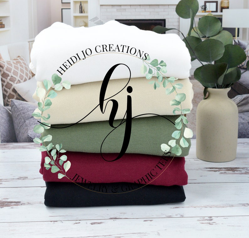 May include: Stack of five blank t-shirts in white, beige, green, burgundy, and black. The shirts are folded and stacked on a white wooden surface. A gold circle with the text "HEIDIJJO CREATIONS" and "JEWELRY & GRAPHIC TEES" is visible in the center of the image.