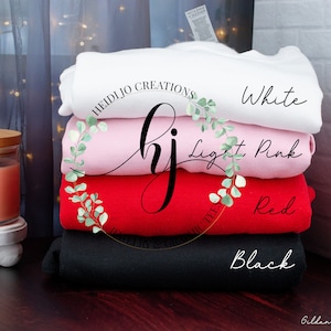 Puede incluir: Pila de cuatro sudaderas en blanco, rosa claro, rojo y negro. Las sudaderas están dobladas y apiladas una encima de la otra. El texto "HeidiJo Creations" es visible en la sudadera de arriba.